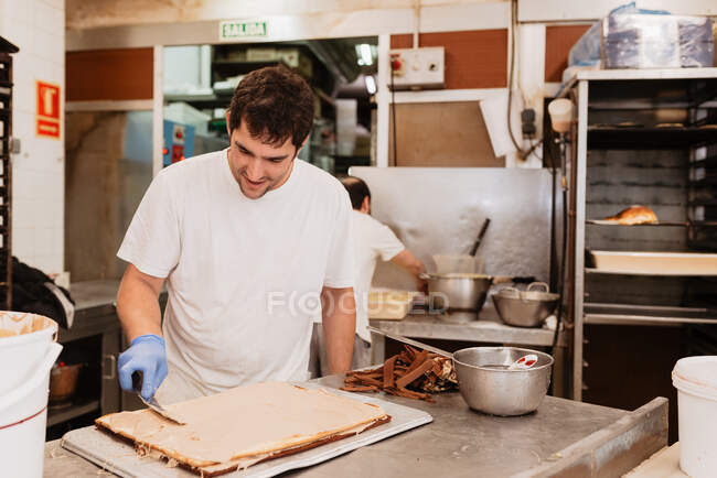 Uomo sorridente in uniforme bianca e guanti in lattice che spalma crema sulla base della torta mentre lavora in cucina — Foto stock