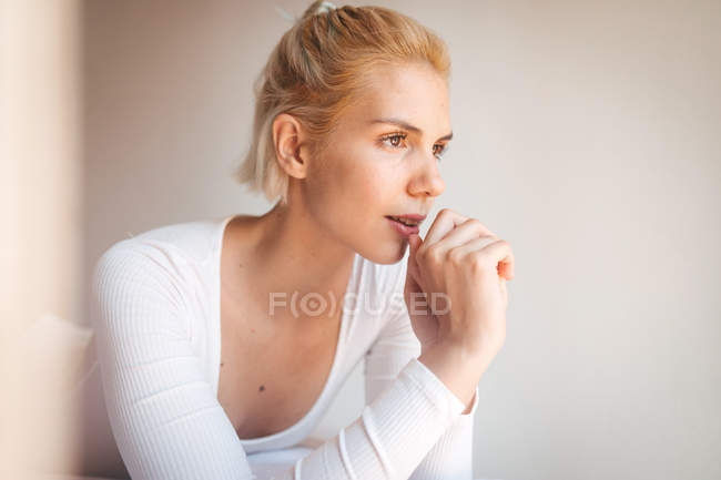 Молодая женщина с светлыми волосами и в телесном костюме, глядя в сторону, сидя на мягкой кровати против белой стены дома — стоковое фото
