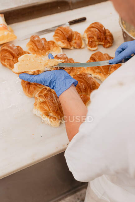 D'en haut travailleur de boulangerie anonyme en gants de latex répandant de la confiture sucrée sur un pain frais sur le comptoir de la cuisine — Photo de stock