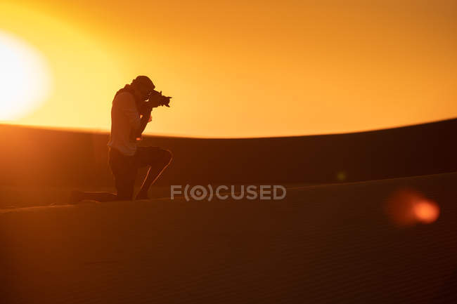 Una silueta de viajero irreconocible que toma fotos de dunas mientras se apoya en la arena en un desierto asombroso. - foto de stock