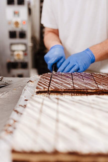 Crop man in uniforme e guanti con coltello per tagliare dolce torta fresca sul tavolo in panetteria — Foto stock