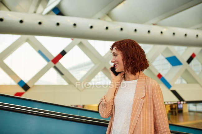Усміхнена молода жінка розмовляє по телефону на вокзалі — стокове фото