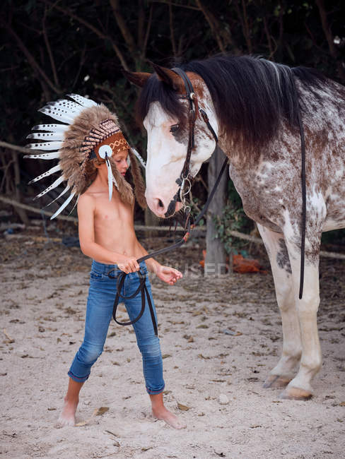 Delighted kid in Indian feather war bonnet with horse on ranch — Stock Photo