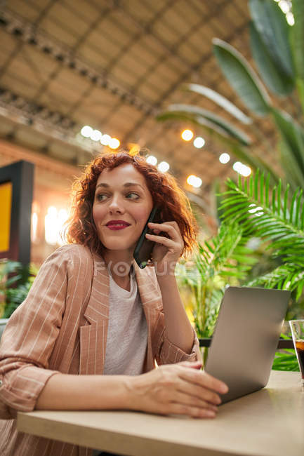 Jovem alegre trabalhando no laptop no café e conversando por telefone — Fotografia de Stock