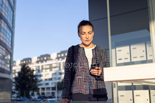 Весела жінка-підприємець в стильній куртці посміхається і використовує смартфон — стокове фото