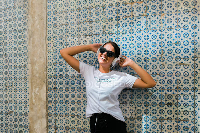 Bella donna spensierata in abito casual e cuffie con le mani alzate accanto al muro di mosaico blu di costruire sulla strada della città — Foto stock