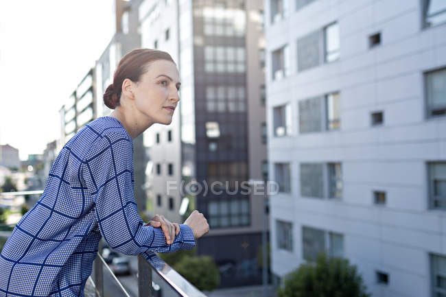 Вид збоку жінки-менеджера в картатій сорочці, посміхаючись і дивлячись геть, стоячи на балконі офісної будівлі — стокове фото
