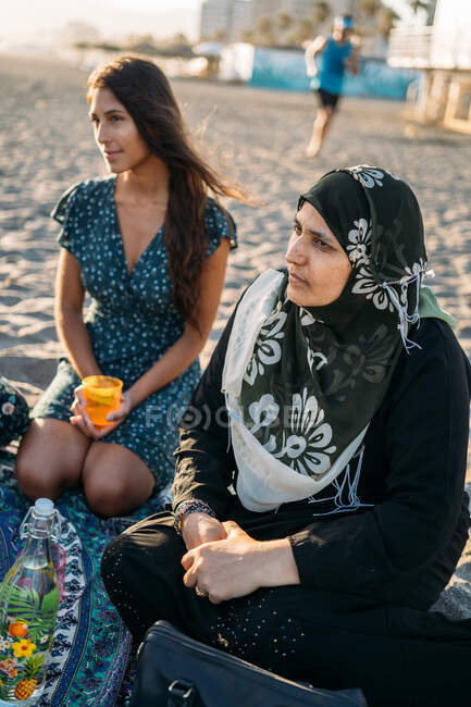 Portrait of two women, one from the Muslim religion and the other girl with brown hair, thoughtful on the beach with her group of friends — Stock Photo