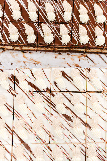 Верхний вид кусочков вкусных белых и коричневых шоколадных тортов, украшенных сливками и соусом на столе в пекарне — стоковое фото