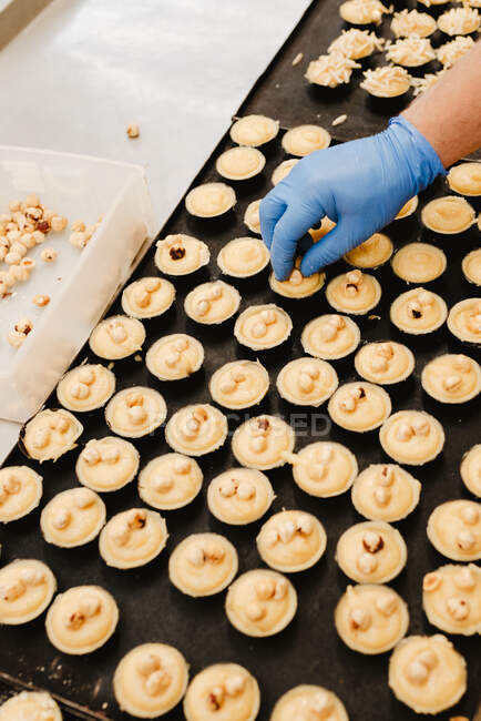 Von oben anonymer Mitarbeiter im Handschuh, der frische Haselnüsse auf leckeres süßes Gebäck auf Tablett legt, während er in der Bäckerei arbeitet — Stockfoto
