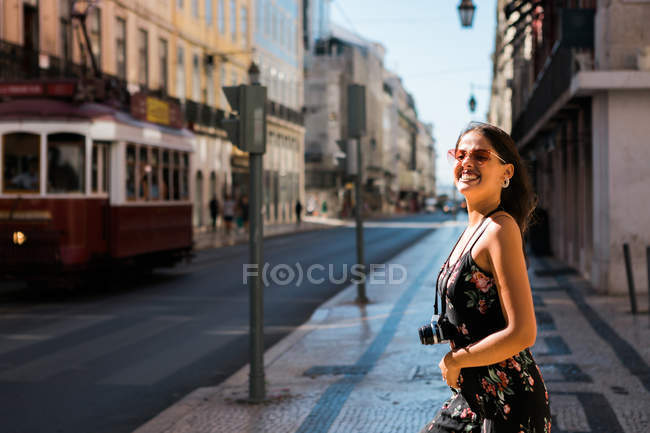 Mujer hermosa y segura en vestido de verano sosteniendo una cámara de fotos mientras está de pie en la calle panorámica de la ciudad soleada en Lisboa, Portugal - foto de stock