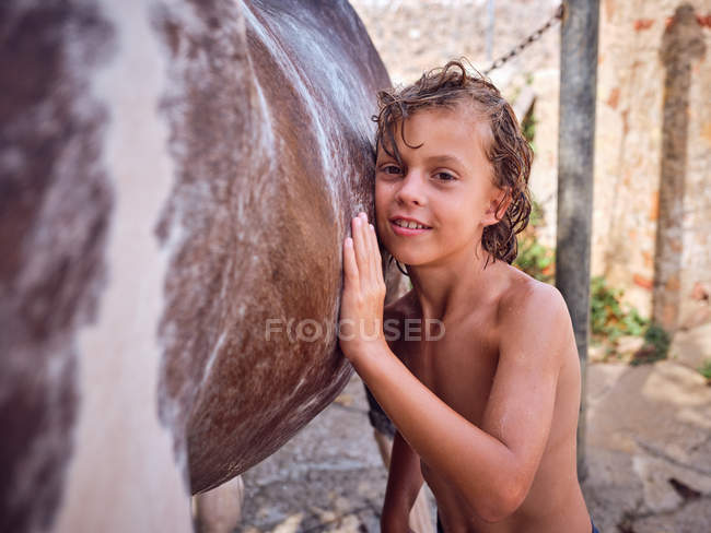 Happy shirtless kid with curly wet hair embracing horse side — Stock Photo