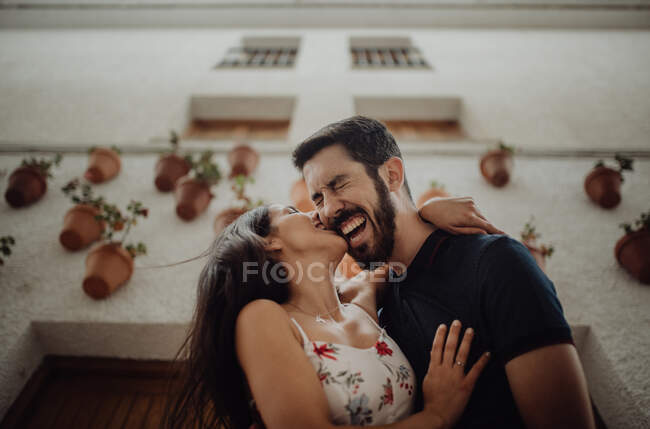 Joyful couple kissing and laughing while standing at street beside building — Stock Photo