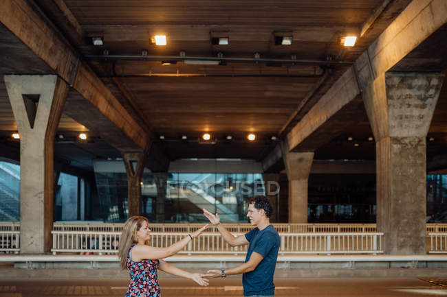Side view of lovely couple spending time together and playing clasping hands game under large bridge in evening lights on city road — Stock Photo