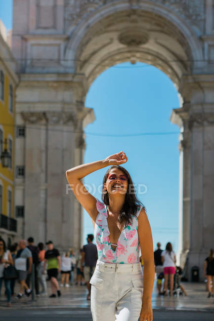 Joven mujer feliz en gafas de sol de pie junto al majestuoso arco en la calle de la ciudad en Lisboa, Portugal - foto de stock