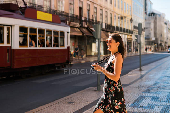 Mujer hermosa y segura en vestido de verano sosteniendo una cámara de fotos mientras está de pie en la calle panorámica de la ciudad soleada en Lisboa, Portugal - foto de stock