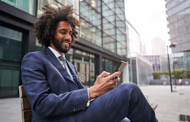 Feliz funcionário afro-americano sentado no banco e sorrindo enquanto navega nas mídias sociais usando smartphone — Fotografia de Stock