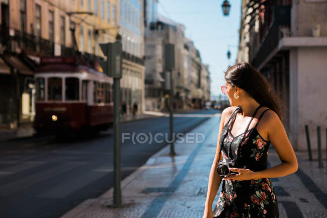 Mulher bonita confiante em vestido de verão segurando uma câmera fotográfica enquanto está em pé na pitoresca rua ensolarada da cidade em Lisboa, Portugal — Fotografia de Stock