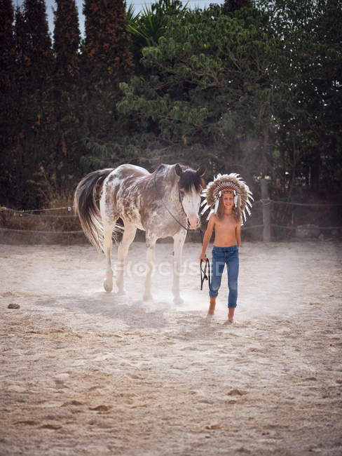 Calm kid wearing feather Indian war bonnet and walking shirtless on sandy farm, leading horse behind — Stock Photo