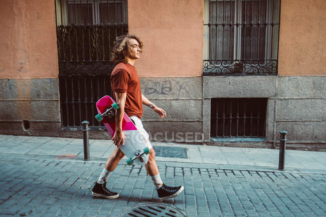 Vue latérale du bel homme sportif tenant longboard tout en marchant sur la ville d'été rue de la ville — Photo de stock