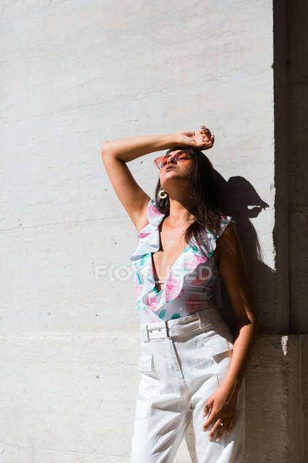 Спокійна розкішна жінка в модному вбранні і блискучі сонцезахисні окуляри, що стоять на білій стіні з руками вгору на мальовничій вулиці — стокове фото