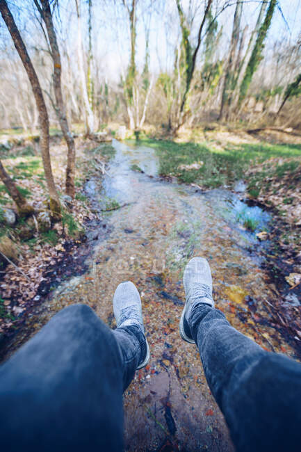 Crop legs of modern male traveler resting and sitting on bridge over shallow water stream in leafless forest on autumn sunny day — Stock Photo