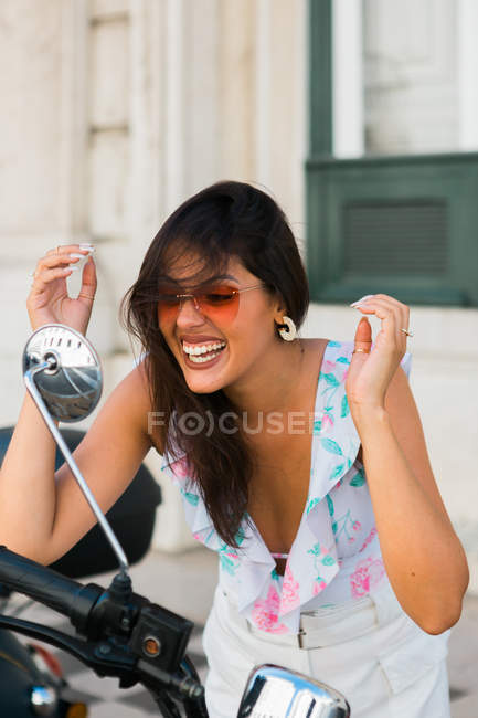 Confident trendy woman grooming hairstyle while standing and admiring self in motorbike mirror on street — Stock Photo
