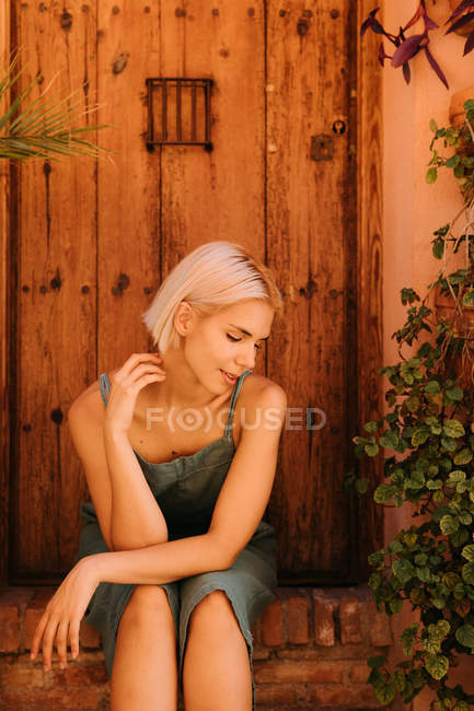 Mujer joven con los ojos cerrados sentado cerca de la puerta de madera y plantas en el patio - foto de stock