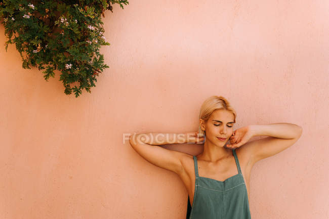 Hermosa joven hembra con el pelo corto y rubio cerrando los ojos y apoyándose en la pared mientras está de pie sobre un fondo rosa borroso - foto de stock