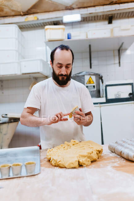 Uomo barbuto in t-shirt bianca mettere pasta fresca in tazze mentre fa pasticceria in cucina di panetteria — Foto stock