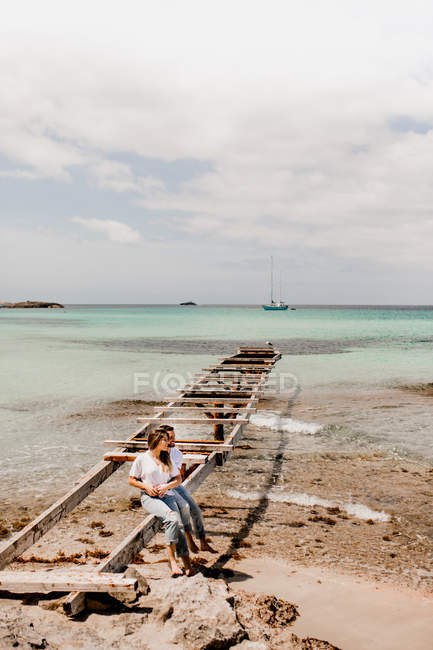 Gli amanti felici seduti sul molo distrutto sulla riva del mare — Foto stock