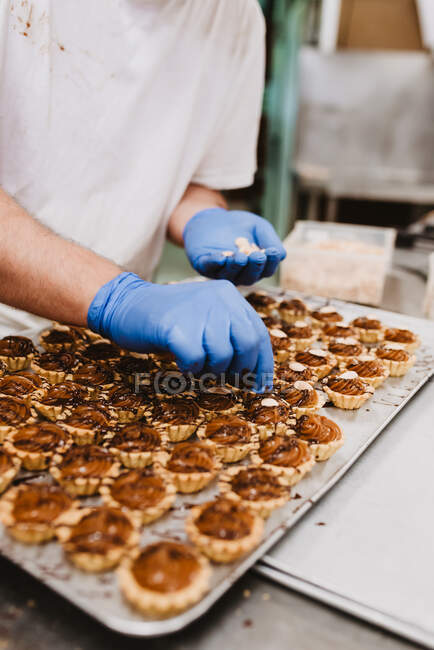 Homme méconnaissable dans des gants en latex mettre des noix sur le dessus de petits gâteaux sucrés tout en travaillant dans la boulangerie — Photo de stock
