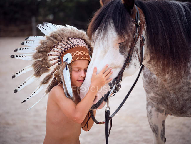 Bambino tranquillo con gli occhi chiusi che indossa tradizionale cappello da guerra indiano, legame con stallone cavallo su sfondo sfocato — Foto stock