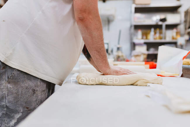 Vista lateral del hombre con sobrepeso de la cosecha en uniforme amasar masa blanda en la mesa mientras se trabaja en la cocina de panadería - foto de stock