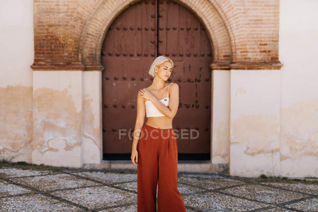 Attractive young woman in stylish outfit folding arms and closing eyes while standing against ancient building — Stock Photo