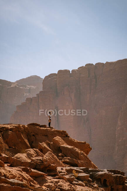 Vista lateral do viajante distante fêmea em pé em penhasco rochoso contra cume da montanha no dia ensolarado no deserto de Wadi Rum — Fotografia de Stock