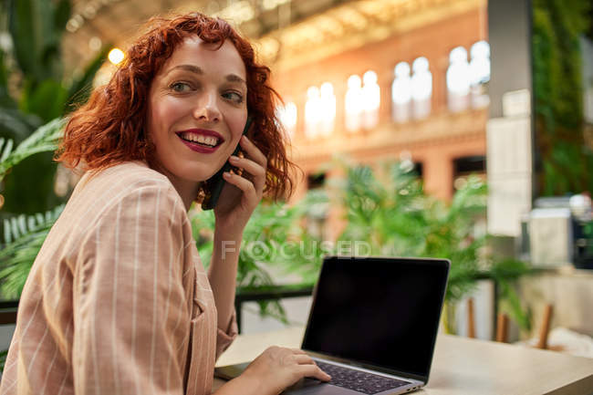 Весела молода жінка працює на ноутбуці в кафе і розмовляє по телефону — стокове фото