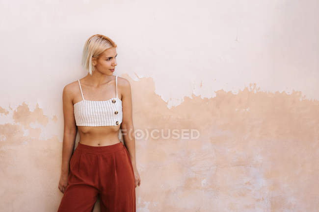 Молода жінка в модному верхній частині і штани посміхаються і дивиться геть, стоячи навпроти стіни будівлі на вулиці — стокове фото