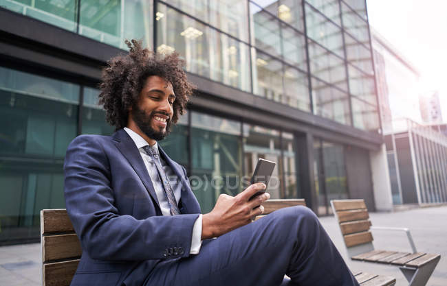 Gioioso uomo africano americano in abito seduto fuori e messaggistica — Foto stock