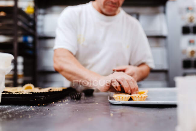 Primer plano empleado anónimo en guantes apretando crema en la parte superior de pasteles de chocolate fresco en bandeja en la panadería - foto de stock