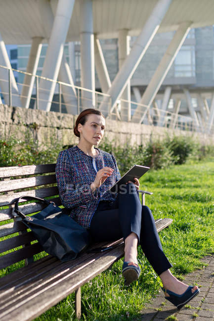 Empresária séria em elegante casaco franzindo a testa e usando tablet enquanto se senta no banco no dia ensolarado no parque da cidade — Fotografia de Stock