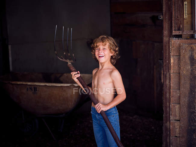 Sério menino sem camisa com forquilha de pé na parede da barraca na fazenda e olhando para a câmera — Fotografia de Stock