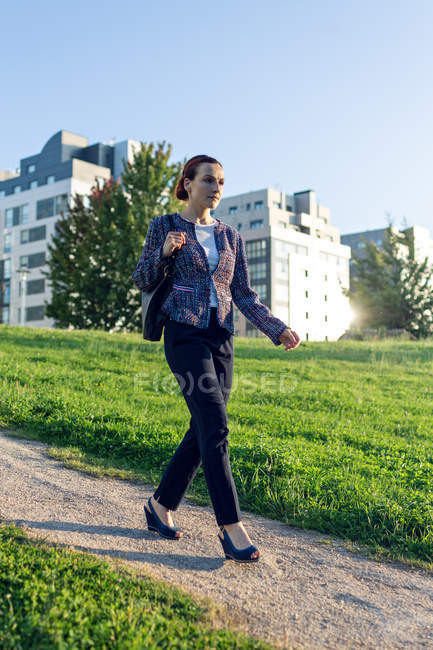Mujer de negocios de larga duración con bolsa caminando por el camino cerca del césped verde en el día soleado en el parque de la ciudad - foto de stock