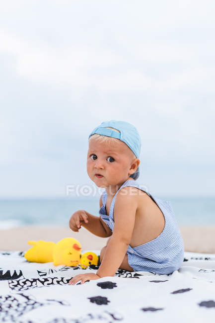 Veduta posteriore di un bambino con un berretto sulla spiaggia accanto alle sue anatre di gomma — Foto stock
