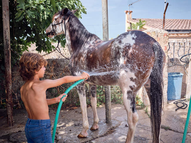 Visão traseira do menino descalço mangueira garanhão com água doce no terraço da fazenda — Fotografia de Stock