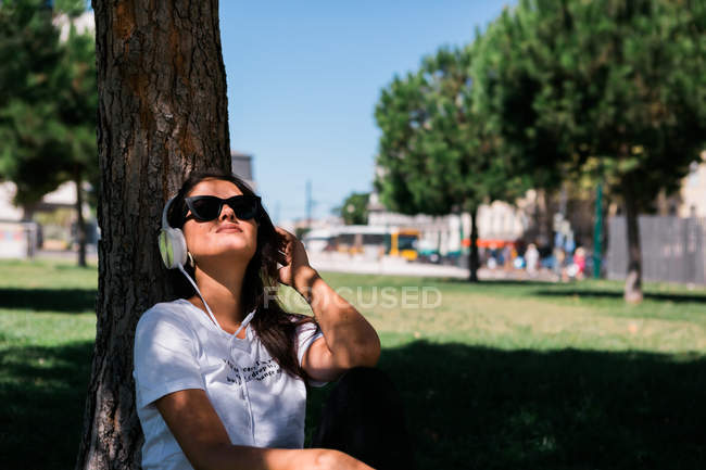 Мирная женщина в солнечных очках и наушниках слушает музыку, сидя на траве в парке — стоковое фото