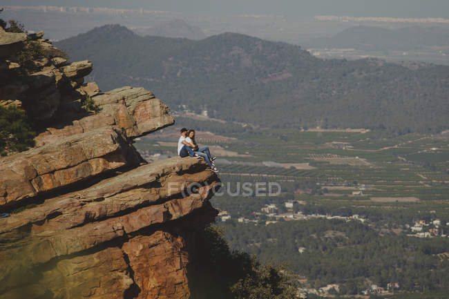 Вид збоку романтичної розслабленої пари в поєднанні вбрання обіймає, сидячи на краю величезної скелястої гори — стокове фото