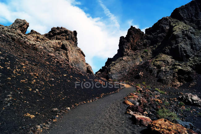 Route étroite et sombre en minéraux volcaniques qui monte jusqu'aux collines rocheuses sous un ciel bleu à Lanzarote, îles Canaries, Espagne — Photo de stock