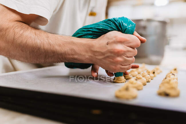 Cocinero anónimo exprimiendo masa de pastelería fresca en bandeja con papel mientras trabaja sobre fondo borroso de panadería - foto de stock