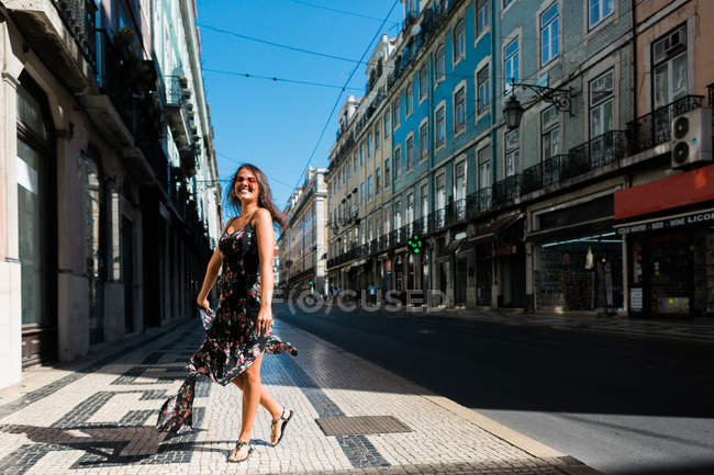 Femme ludique en robe longue se retournant tout en marchant le long de la rue d'été de la ville — Photo de stock